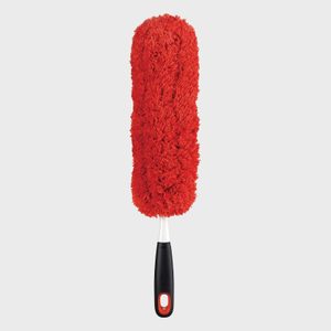 Red fluffy duster rests vertically with a black handle, against a plain light gray background, suitable for cleaning surfaces.