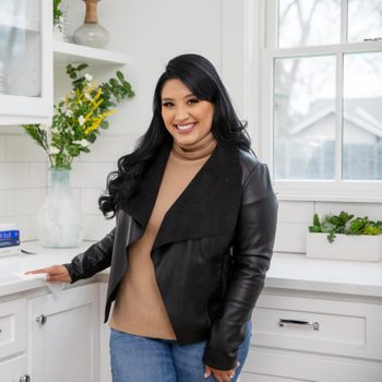 A person smiles while standing in a bright kitchen, wearing a black leather jacket and holding a paper towel, with plants in the background.