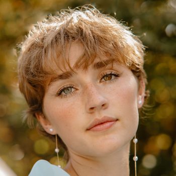 Person gazing softly, set against a blurred backdrop of greenery with warm sunlight enhancing their hair and complexion.