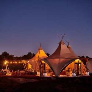 Illuminated tent structures glow softly with warm lights, surrounded by outdoor seating and string lights, under a clear twilight sky in a natural setting.