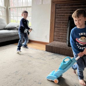 Two children play; one vacuums with a toy, the other mimics guitar, inside a living room with a couch and large windows.