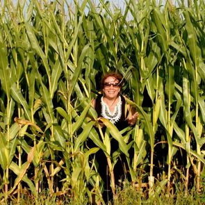 A person stands smiling, emerging from tall green corn stalks under a clear blue sky.