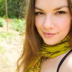 A woman smiling, wearing a patterned yellow scarf, with blurred greenery in the background.