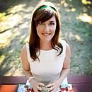 A woman smiles while sitting at an outdoor table with a floral tablecloth, surrounded by grass and dappled sunlight.