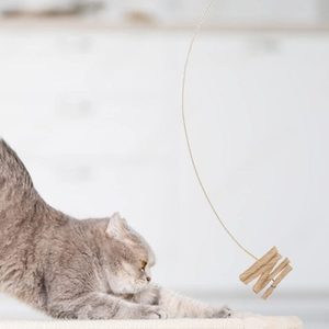 Cat stretches on a soft surface, focusing on a dangling wooden toy. Background features a blurred, bright interior setting.