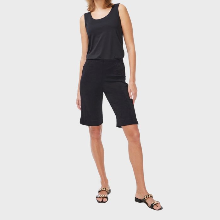 Woman wearing black tank top and shorts stands confidently, modeling black sandals with gold chain detail. Plain white background emphasizes her attire.