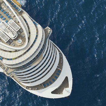 Cruise ship sails on open ocean; aerial view shows decks with pool and loungers, surrounded by deep blue water.