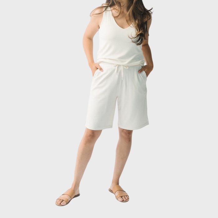 Person stands, hands in pockets, wearing a sleeveless top and shorts, with sandals, against a plain background.