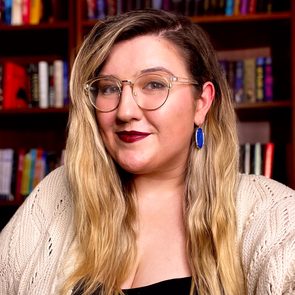 A person with long hair and glasses smiles, wearing a sweater, in front of a bookshelf with colorful books.