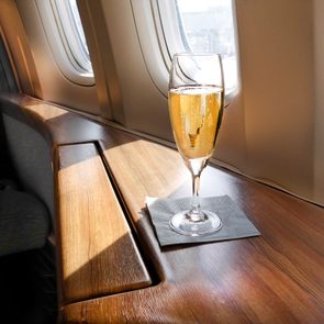 A champagne glass sits on a wooden airplane tray table, bathed in sunlight streaming through nearby windows.