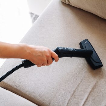 Vacuum nozzle cleans beige couch; hand grips handle; modern, tidy room.