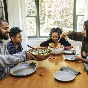 Family shares salad and bread at a dining table, surrounded by windows and a plant, creating a warm, homely atmosphere.