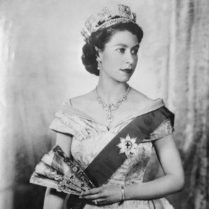 A woman wearing a crown and elegant gown holds a decorative fan, standing in a formal setting with a textured curtain backdrop.