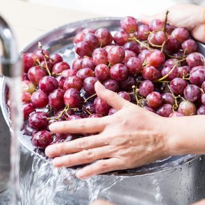 Hands wash red grapes in a stainless steel bowl under a running faucet in a kitchen sink.