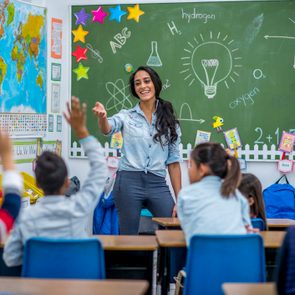 Teacher engaging students, raising hands eagerly in a colorful classroom with a world map and scientific drawings on the chalkboard.