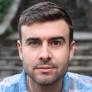 A person looks directly at the camera, wearing a plaid shirt, with stone steps blurred in the background.
