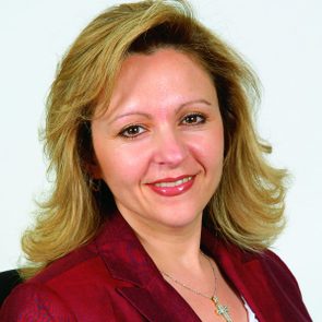 Smiling woman wearing a red blazer and cross necklace, set against a plain, light background.