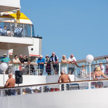 People gather on a ship deck, enjoying a sunny day, surrounded by railings and life boats. Some lounge, while others take photos.