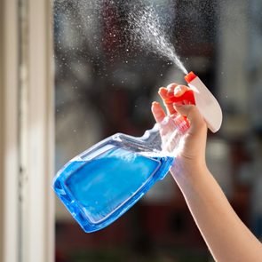 Hand sprays blue liquid from a plastic spray bottle indoors near a window, with droplets visible in the air.