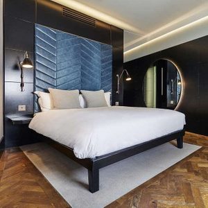 Bed sits neatly, adorned with white linens and pillows, against a dark, modern panel wall in a stylishly lit room.