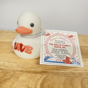 Duck sits on a wooden surface, displaying a note about a Royal Caribbean cruise and Mills family, adorned with text and decorative hearts.