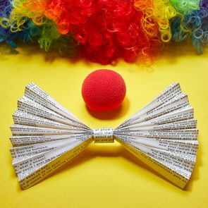 Paper bow tie with text rests beside a red foam nose, beneath a multicolored curly wig on a vibrant yellow background.