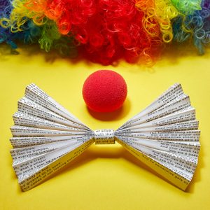 Paper bow tie with text rests beside a red foam nose, beneath a multicolored curly wig on a vibrant yellow background.
