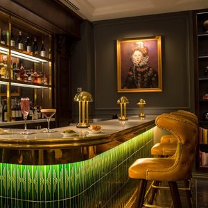 Bar counter displays cocktails, plates; three lamps illuminate, surrounded by green-tiled front. Elegant chairs line bar beneath portrait of a regal figure.