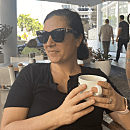 Person holding a mug, sitting at an outdoor cafÃ©, wearing sunglasses and a dark shirt. In the background, people walk by modern buildings.