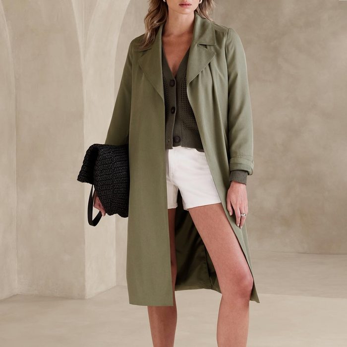 A woman wears a green trench coat and white shorts, holding a black woven handbag against a neutral-toned background.