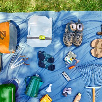 Camping gear arranged on a blue tarp, surrounded by grass; includes backpacking tent, water jug, hiking boots, sandals, camera, utensils, and safety equipment.