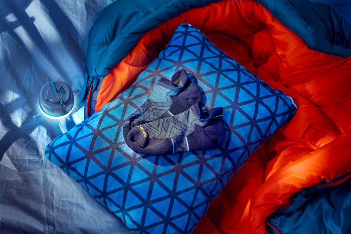 Socks rest on a patterned blue pillow beside an opened blue and orange sleeping bag on a bed, with a small light nearby.