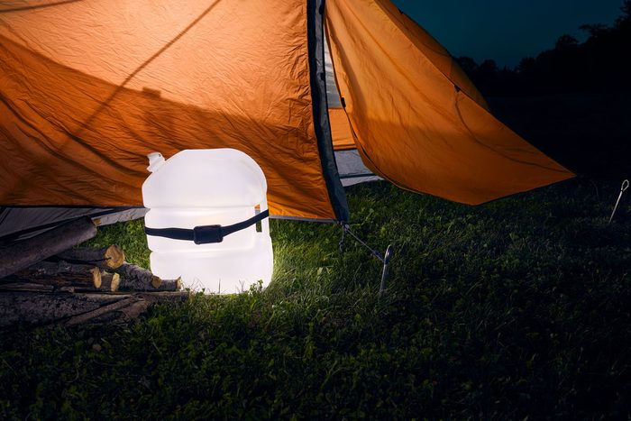 A translucent water container glows beside chopped firewood near an orange camping tent on grassy ground at night.