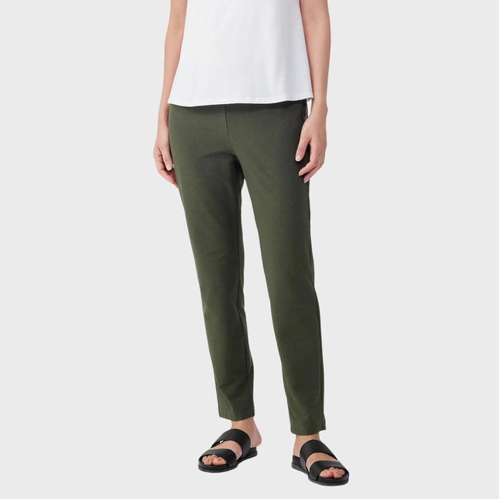 Person standing, wearing olive green pants and a white top, with black sandals against a plain, light grey background.