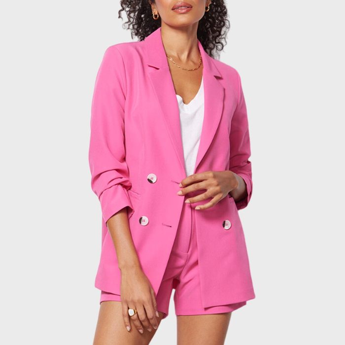 A person stands wearing a bright pink blazer and shorts, hands adjusting the blazer. Background is plain white.