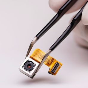 Tweezers hold a small camera module with an attached flex cable in a close-up view, set against a neutral background.