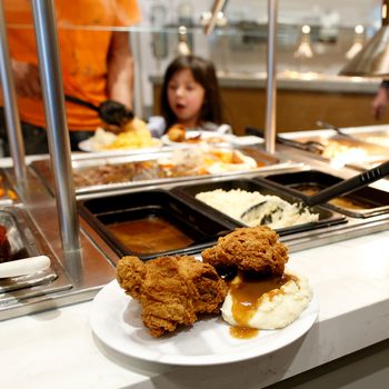 Plate with fried chicken and mashed potatoes sits at a buffet counter, surrounded by various food trays, including meatloaf and vegetables.