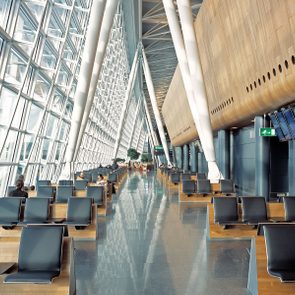 Seats sit empty in a modern, spacious airport terminal with large windows and sleek design, creating an airy, open atmosphere.