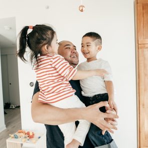A person holds two smiling children inside a bright room with wooden cabinets and a small toy shelf.
