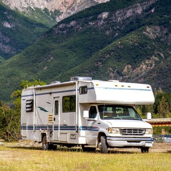 A white RV is parked on a grassy area, surrounded by lush green mountains and trees near a small bridge over a stream.