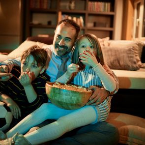 A family sits on the floor watching television, eating popcorn, and surrounding a cozy living room with bookshelves and a couch.