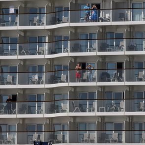 Multiple people stand on cruise ship balconies, observing or relaxing, surrounded by white railing and blue-tinted glass doors, with patio furniture visible.