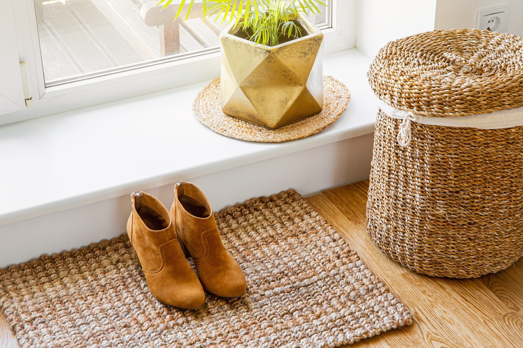 Hardwood floor with jute doormat, shoes and flower pot and seagrass laundry basket by window. Natural material objects in home concept. Home interior.