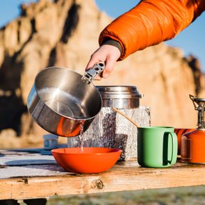 Person pours liquid from a metal pot into a red bowl on a wooden table, outdoors with rocky cliffs in the background.