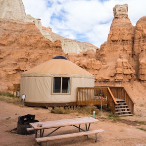 A tan yurt sits beside a wooden deck nestled among rocky, reddish cliff formations under a partly cloudy sky. A picnic table and fire pit nearby.