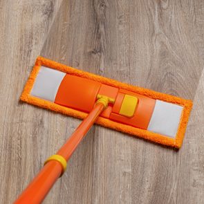 Mop moving across wooden floor, cleaning surface with an orange microfiber pad, leaving a visible clean stripe behind.