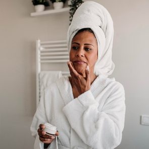 Woman applies cream to face, standing in a bathroom, wearing a bathrobe with a towel wrapped around her head.