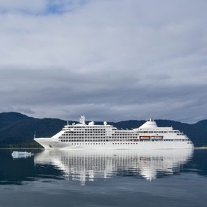 Cruise ship sails calmly on reflective water, surrounded by evergreen hills under a cloudy sky.