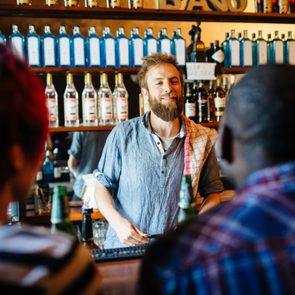 Bartender smiles behind the bar, facing two patrons. Shelves of liquor bottles line the wall, creating a lively and inviting atmosphere.