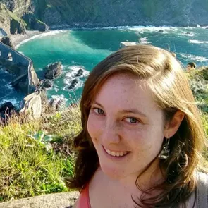 A person smiles outdoors, with coastal cliffs, turquoise water, and a winding stone path in the background.
